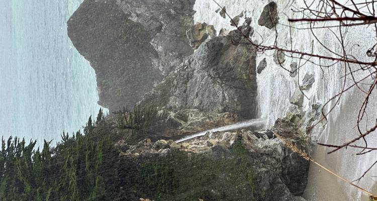 Vertical view of a slender waterfall cascading from lush cliffs to a small beach cove.