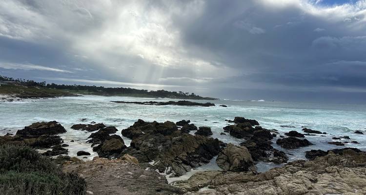 Sunbeams breaking through dramatic clouds over a rocky Pacific shoreline and crashing surf.