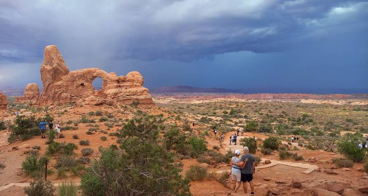 Visitors scattered across viewpoint overlooking a large sandstone arch formation before dark storm clouds