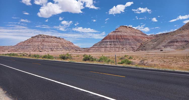 Freshly paved blacktop highway flanked by striped desert buttes under a bright blue sky.