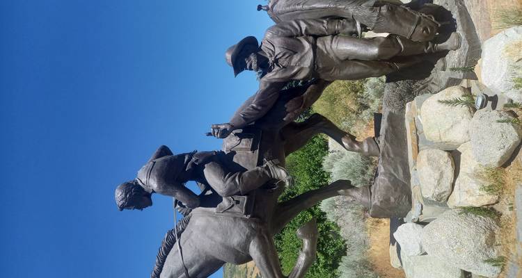 Bronze statue group depicting a mounted rider handing off mail, set against clear blue sky and shrubs.