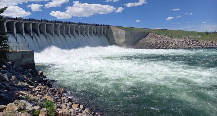 Mächtiger Wasserstrom, der von einem Betondamm in den darunter tobenden Fluss unter blauem Himmel hinabstürzt.