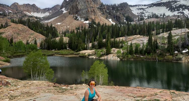 Smiling hiker in sunglasses posing beside a calm mountain lake surrounded by evergreens and rocky peaks.