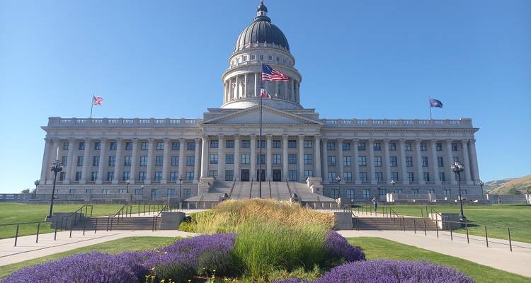 Grand neoclassical Utah State Capitol with flying flags and purple flower beds leading to its steps.