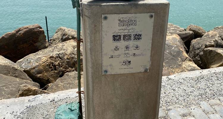 Close-up of a worn informational plaque mounted on a concrete post beside coastal rocks and turquoise water.