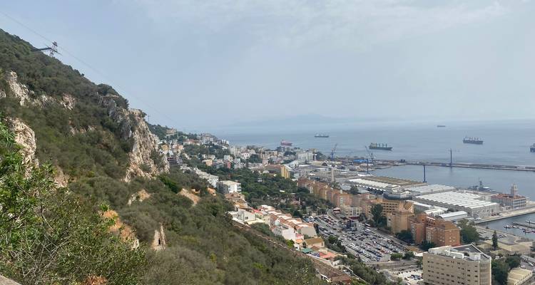 High vantage of Gibraltar town, harbour and shipping lanes with cable-car line descending the green slope.