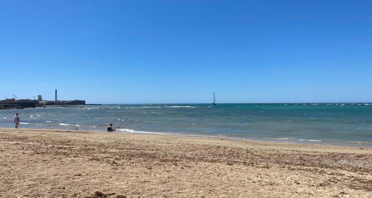 Wide sandy beach with shallow waves, a distant lighthouse and a couple of bathers under a clear blue sky.