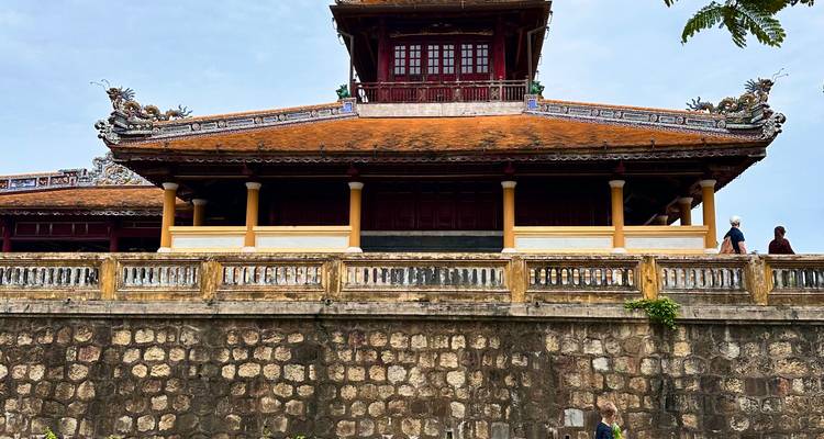 Ein langes kaiserliches Pavilion mit orangefarbenem Dach sitzt auf einer Steinmauer, während Besucher entlang der Wehrgänge spazieren.