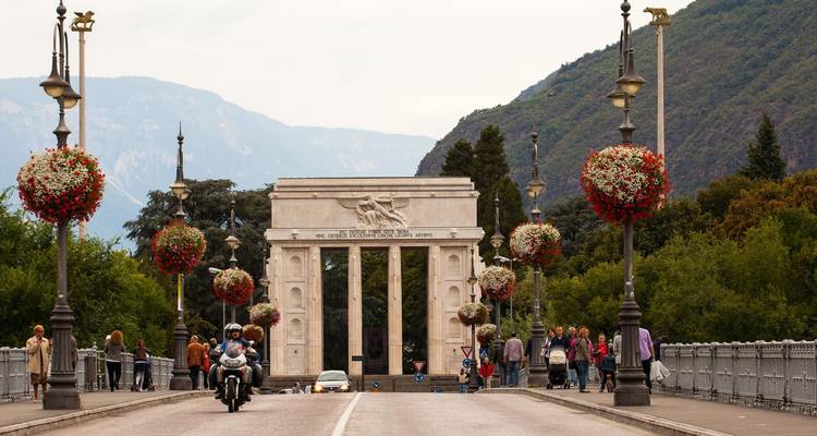 Stone bridge leading to a neoclassical triumphal arch framed by mountains and floral lampposts, with pedestrians and a motorbike.