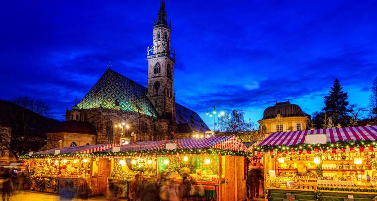 Nighttime Christmas market with striped stalls glowing under fairy lights in front of Bolzano cathedral.