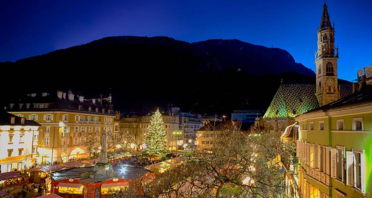 Elevated view over a brightly lit alpine town square with a large Christmas tree and market stalls after dusk.