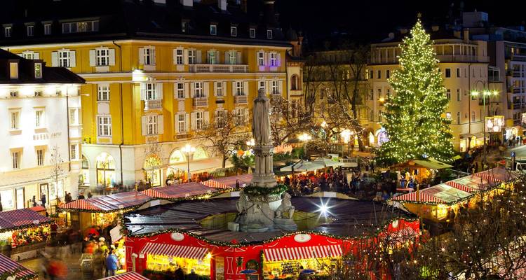Place du marché de Noël animée la nuit avec une fontaine illuminée, des étalages rayés et des foules.