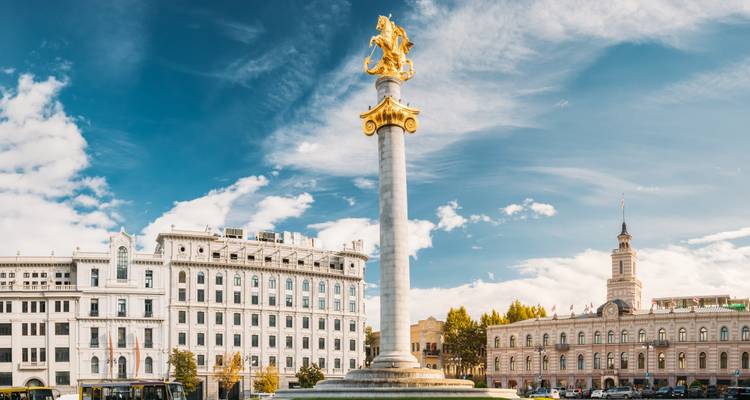 Estatua de San Jorge en lo alto de una columna alta en la Plaza de la Libertad de Tbilisi con cielos azules y fachadas clásicas.