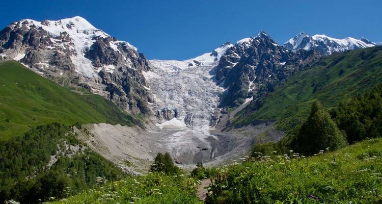 Massive glacier descending between rugged Caucasus mountains under a clear summer sky.