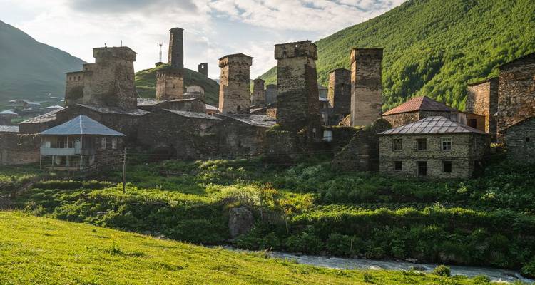 Ancient stone Svan towers rise above green meadows and a stream in a remote Georgian highland village.