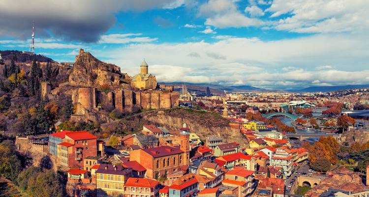Panoramic aerial of Tbilisi old town with Narikala Fortress and colourful rooftops under dramatic clouds.