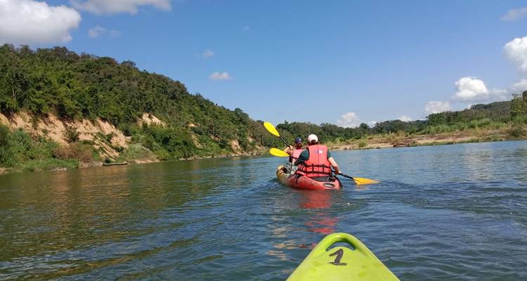 View from a kayak following two paddlers on a calm tropical river flanked by forested banks.
