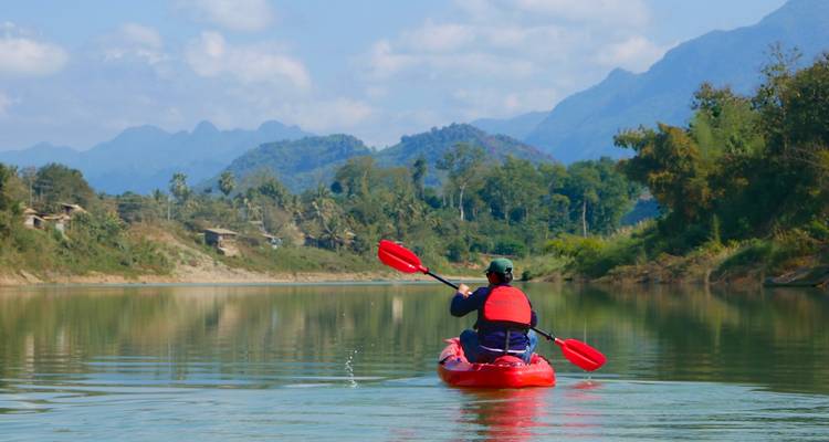 A lone kayaker glides across glassy water toward blue mountains under a clear sky.