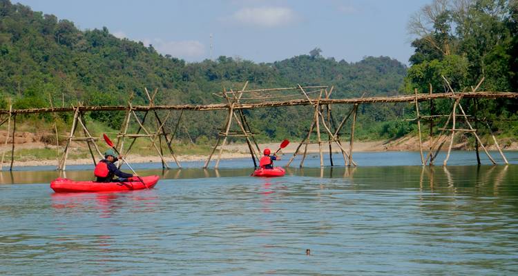 Pair of kayakers approach a rustic wooden river bridge backed by forested hills.