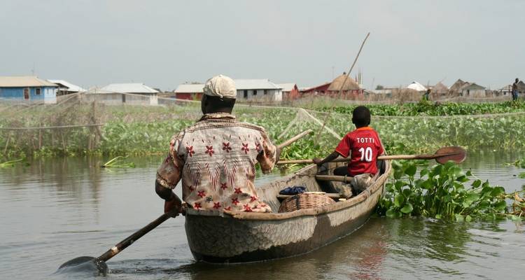 Twee mannen die met peddels een houten uitgehakte kano door waterplanten in een drijvend dorp voortbewegen.