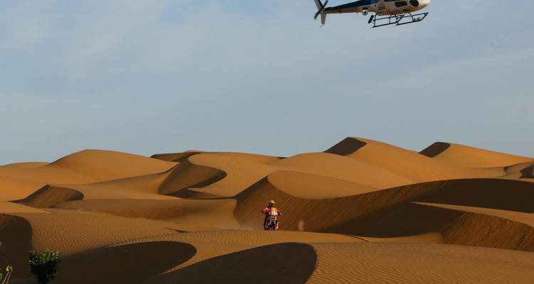 Motorcyclist navigating the rolling dunes of Lompoul Desert with a helicopter above.