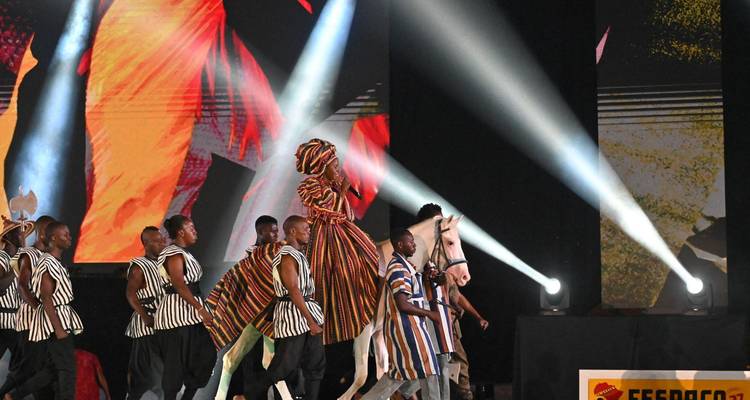 Stage performance with dancers in traditional striped costumes, rider on white horse and dramatic spotlights.