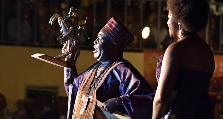 Man in traditional attire proudly holding up a bronze trophy on stage beside a presenter.
