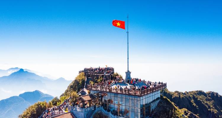 Luftaufnahme des Fansipan-Gipfelkomplexes mit hohem Fahnenmast über einem Wolkenmeer und Bergspitzen.