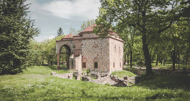 A small medieval stone church surrounded by leafy green parkland and ruins.