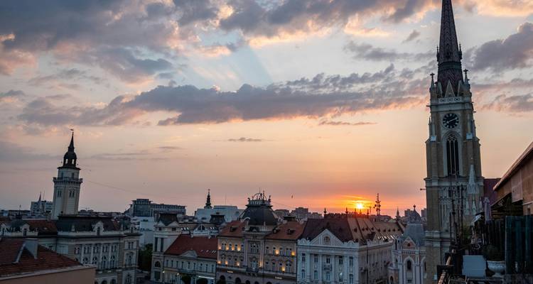 Sunset over the rooftops of Novi Sad with church spires silhouetted against colourful clouds.