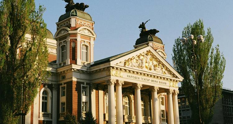 Neoclassical National Theatre in Sofia framed by tall trees under clear evening light.