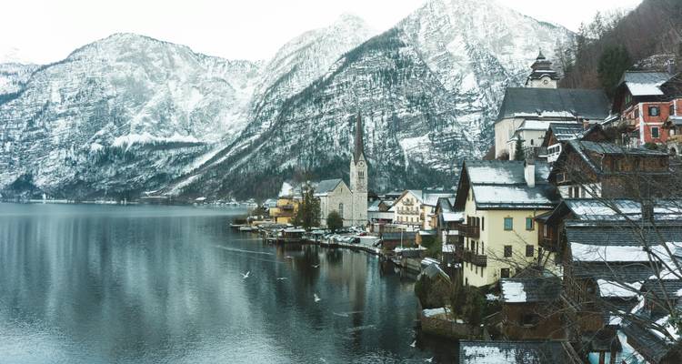 Winter scene of Hallstatt with snow-covered alpine houses reflecting in the calm lake below towering mountains.