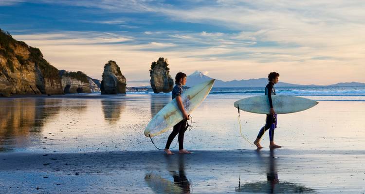 Zwei junge Surfer tragen Bretter entlang eines reflektierenden Strandes mit dramatischen Meeresstapeln unter einem farbenfrohen Abendhimmel