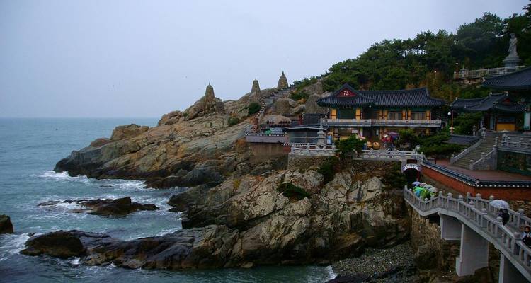 Buddhistische Tempelanlage an der Küste, auf felsigem Ufer gelegen, mit Wellen darunter bei bedecktem Himmel