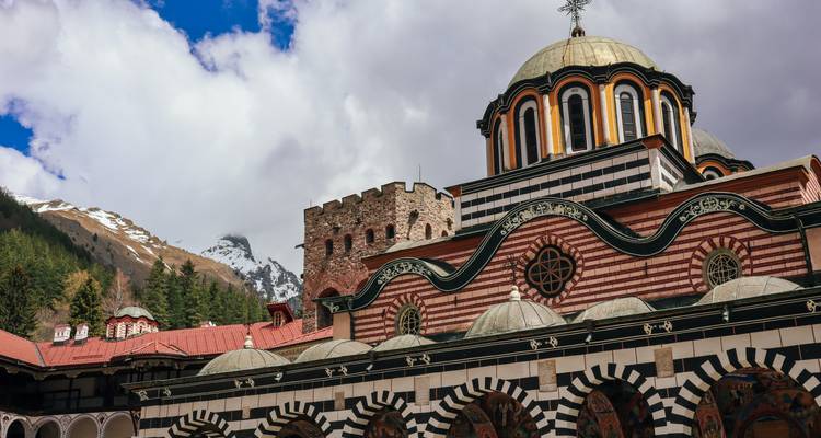 Striped façade and domes of Rila Monastery with snow-capped mountain backdrop