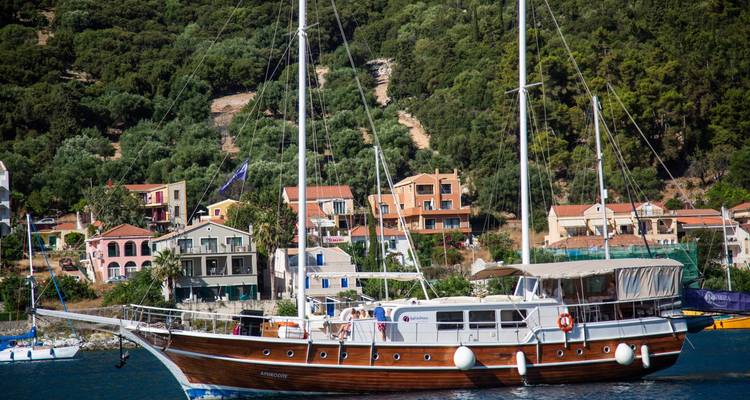 Velero gulet de madera anclado cerca de un pintoresco pueblo costero lleno de colores y rodeado de verdes colinas.