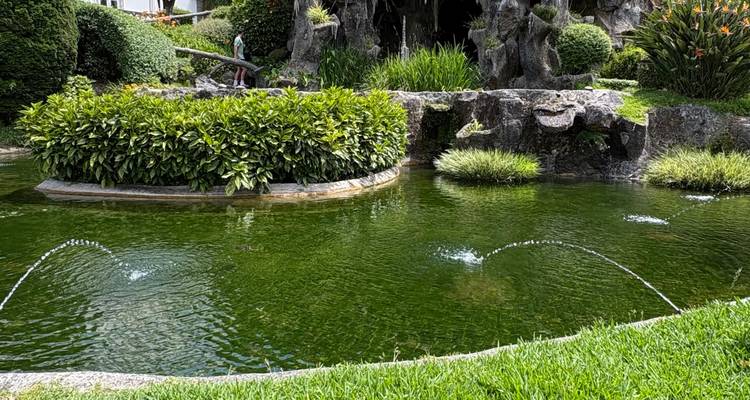 Bassin formel avec de petits jets d'eau et une verdure luxuriante à côté d'une grotte de rocher.