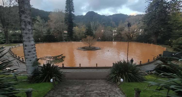 Circular thermal pool filled with rusty-colored mineral water surrounded by gardens and trees.