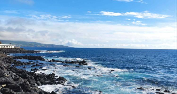 Rocky volcanic coastline with waves crashing under a bright blue sky.