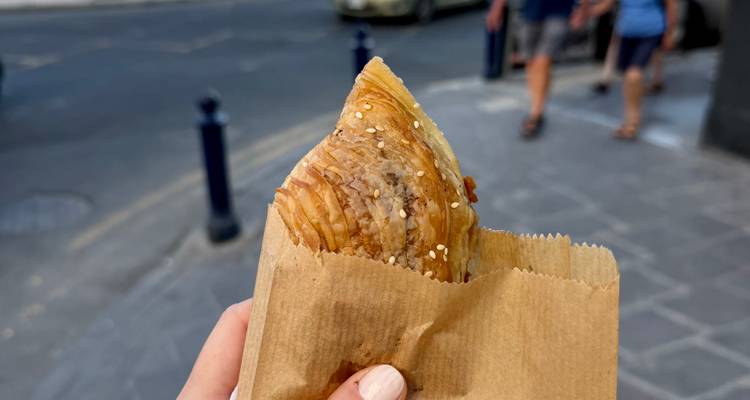 Hand holding a flaky sesame-topped pastry in a paper bag on a Maltese street.