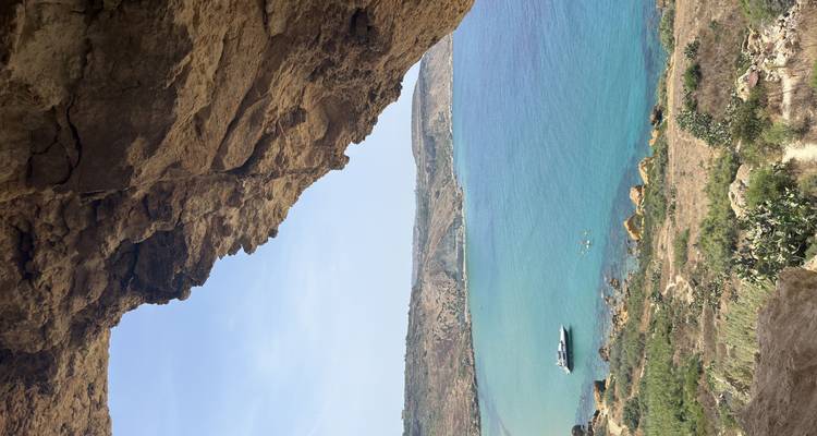 Rocky cave opening framing a tranquil turquoise bay with a small boat below.