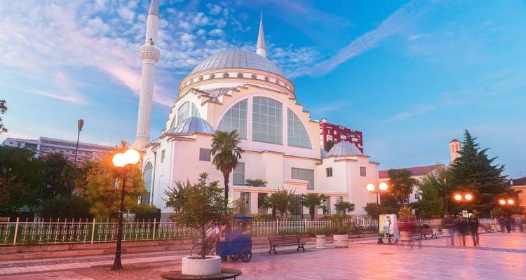 Mosquée blanche moderne avec un haut minaret et des palmiers sur une place urbaine lumineuse.