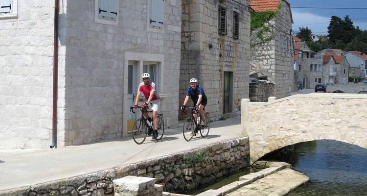 Deux cyclistes sur route traversent un village en pierre en passant par un petit pont en arc et un canal.