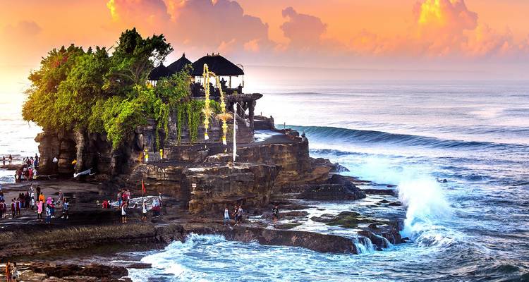Crowds gather around Tanah Lot temple perched on a rocky outcrop amid crashing waves at golden sunset.