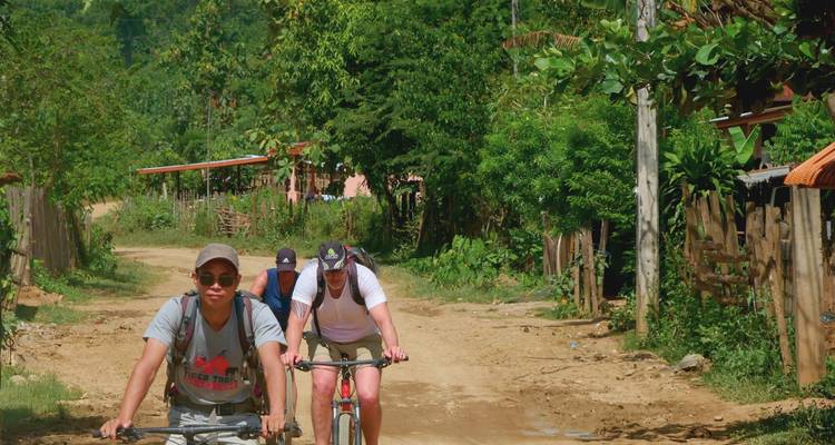Cyclists pedal along a dusty village track lined with tropical greenery in rural Laos.