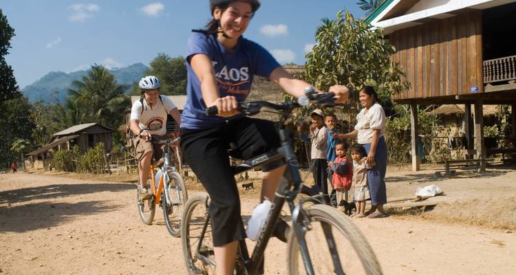 Smiling riders cycle past village children who watch from a dusty roadside in mountainous Laos.