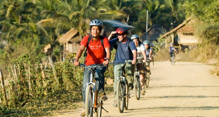 A line of cyclists follows a dirt path flanked by palms and bamboo huts in rural Laos.