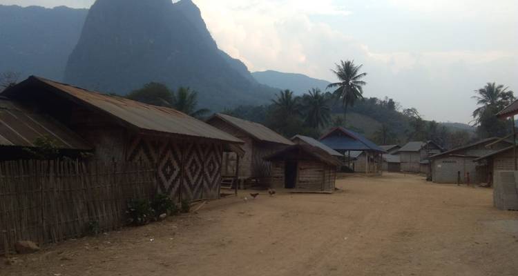 Quiet dirt street lined with wooden stilt houses beneath a towering dark mountain silhouette.