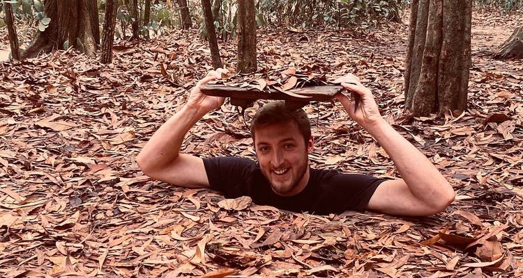 Man emerging from a concealed tunnel hatch in a forest floor covered with dry leaves