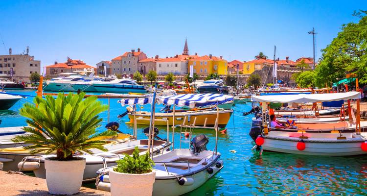 Colorful small boats and luxury yachts moored in a bright turquoise harbour with pastel houses and a church spire beyond